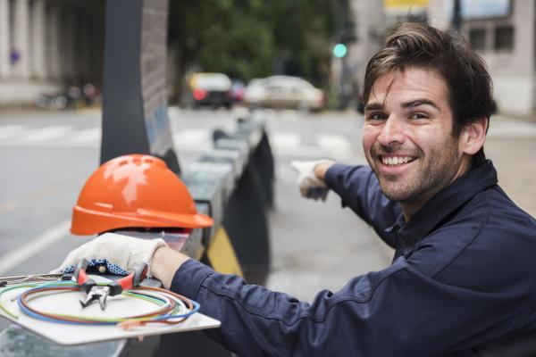 portret van glimlachende mannelijke elektricien die met bouwvakker en materiaal op straat richten